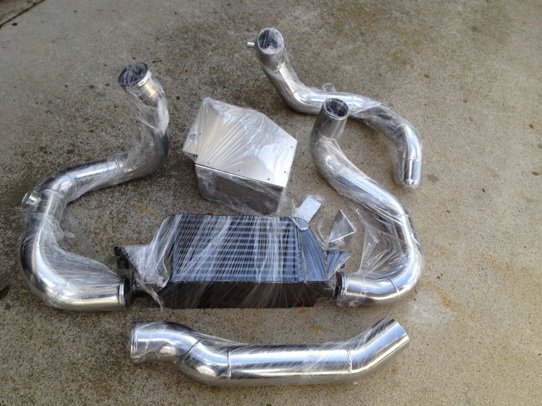Shiny metal pipes and a black intercooler, all wrapped in plastic, laid out on concrete