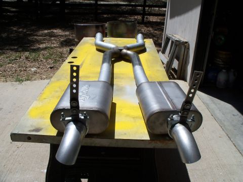 1979 Ford Ranchero - A complete stainless dual exhaust system laid out on a yellow-painted workbench, with an X-pipe crossover, two oval mufflers, angle-cut tip…