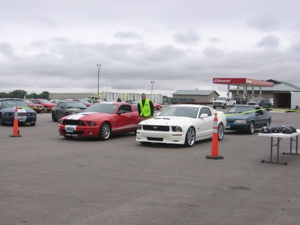 Several Ford Mustangs parked in a lot, with a gas station in the background