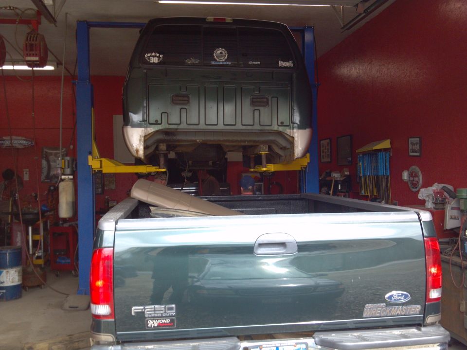 Rear view of a dark green Ford F-250 Super Duty truck, with another truck lifted above it in a garage