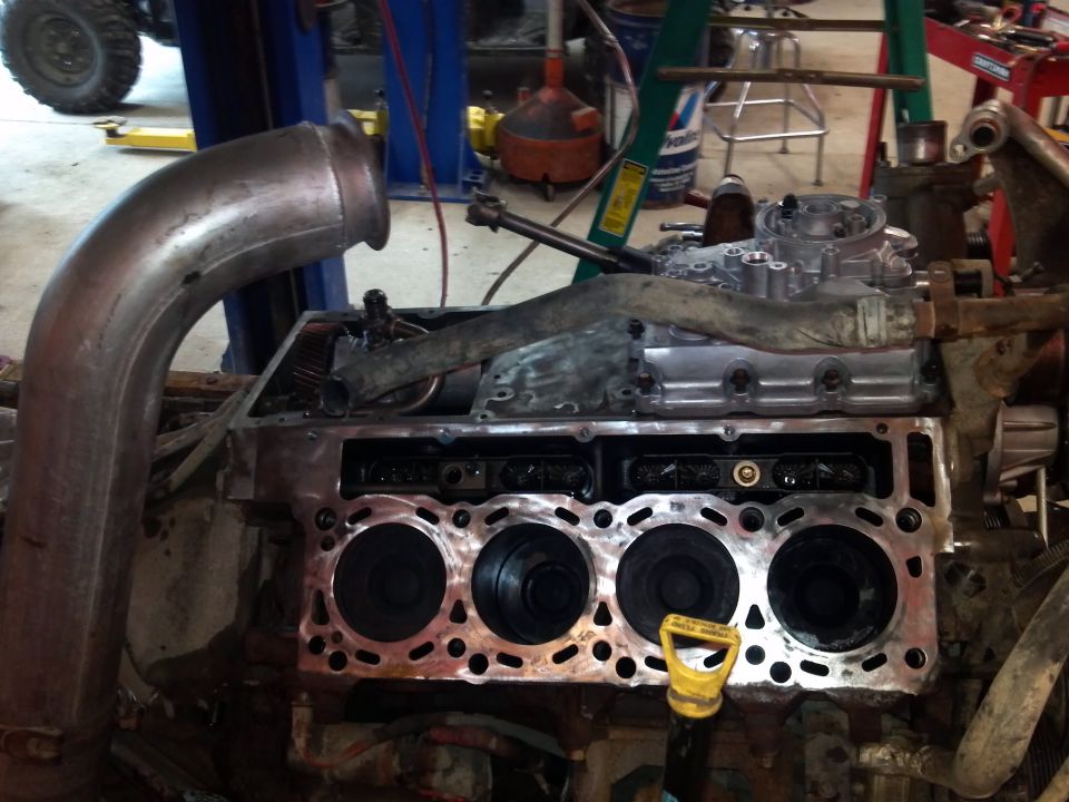 Engine block with cylinders exposed, large metal exhaust pipe on left, yellow dipstick visible