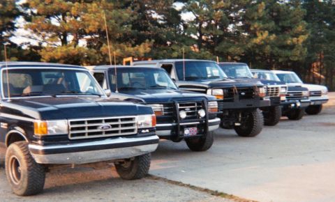 1988 Ford Bronco - A line of Ford Bronco SUVs and trucks are parked in a row, viewed from the front 3/4 angle.