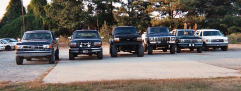 1988 Ford Bronco - A lineup of Ford Bronco SUVs are parked on a paved surface with trees in the background.
