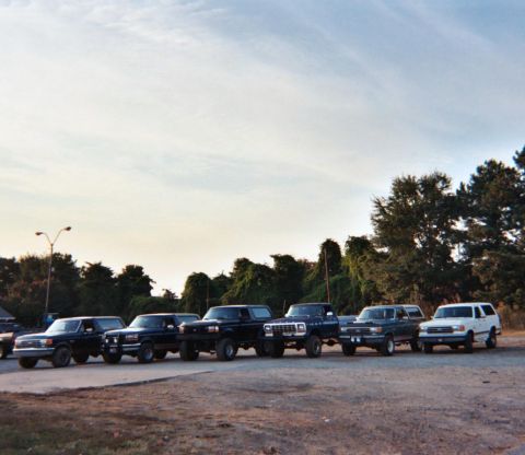1988 Ford Bronco - A group of Ford trucks and SUVs are parked in a row under a cloudy sky.
