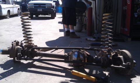 1978 Ford Bronco - Front axle assembly with coil springs and shocks removed, with trucks in the background.