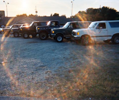 1988 Ford Bronco - Several Ford Broncos are lined up in a parking lot, with the sun setting behind them.