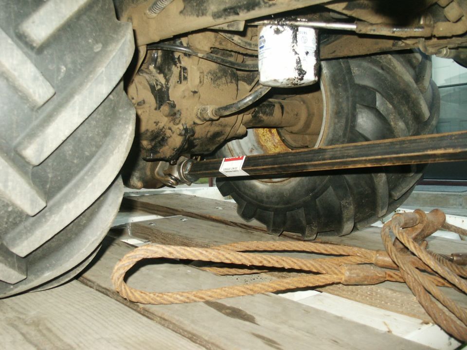 Undercarriage of a tractor showing the drive shaft, oil filter, and large tire. Ropes and wooden planks are in the foreground