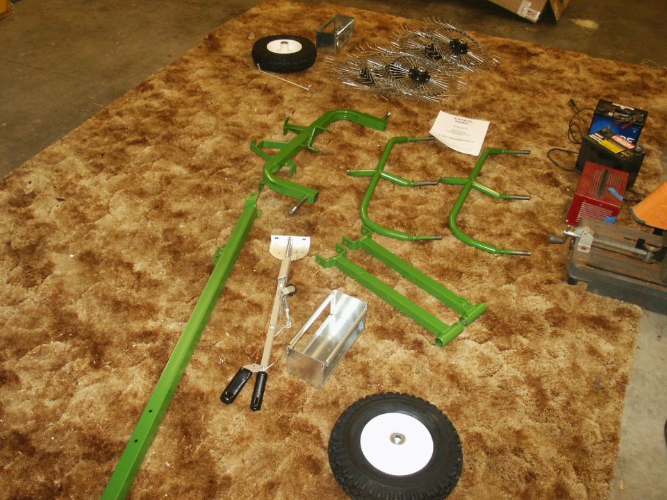 Green metal frame pieces, two wheels, and rake tines laid out on a brown carpet