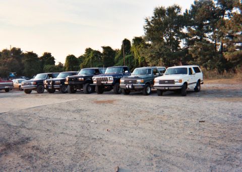 1988 Ford Bronco - Several trucks and SUVs are parked in a gravel lot with trees in the background.