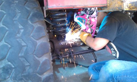 1978 Ford Bronco - A person welds on a vehicle's suspension system, with a large tire visible in the foreground and sparks flying.