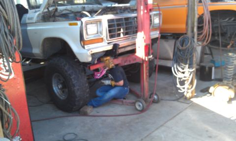1978 Ford Bronco - A mechanic works on the underside of a white 1978 Ford Bronco in a garage, with another vehicle visible in the background.