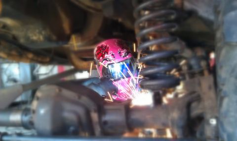 1978 Ford Bronco - A welder in a pink helmet is actively working on a vehicle's suspension system, with sparks flying.