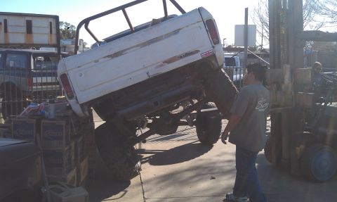 1978 Ford Bronco - A forklift is lifting a white Ford Bronco, showing its undercarriage and rear end.