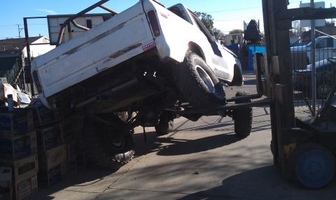 1978 Ford Bronco - A forklift is lifting a white pickup truck, which is precariously balanced on top of stacked boxes.