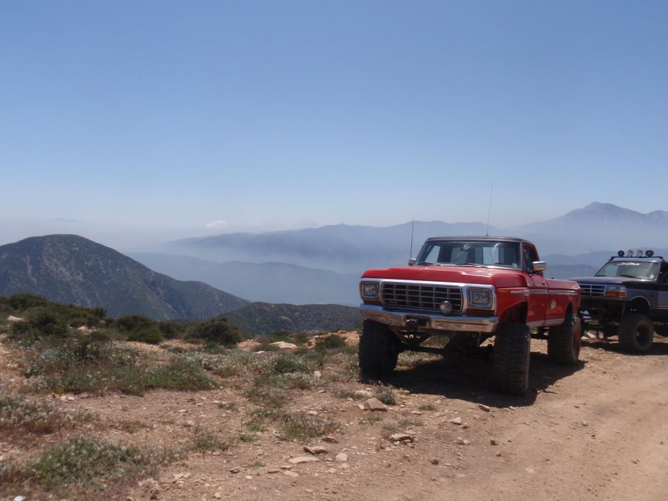 Red 1978 Ford F-150 pickup truck and a dark SUV parked on a dirt road with mountains in the background