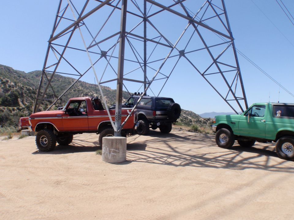 Three Ford Broncos parked under a power line tower. Orange Bronco in foreground, dark blue Bronco behind it, green Bronco to the right
