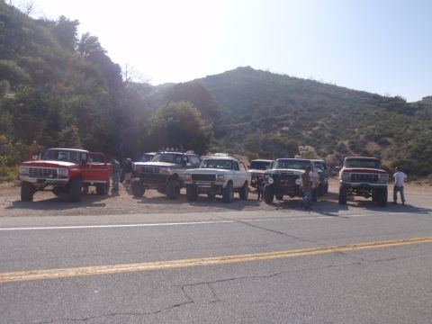 1978 Ford Bronco - A group of modified Ford trucks and SUVs are parked on the side of a road with a mountain range behind them.