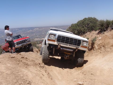 1978 Ford Bronco - Two trucks navigate a steep dirt path on a sunny day, with a person observing from the side.