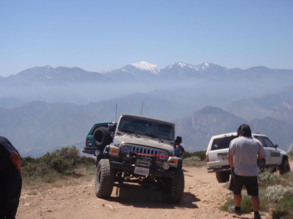 Tan Jeep Wrangler with winch and spare tire on rear, parked on dirt road with mountains in background