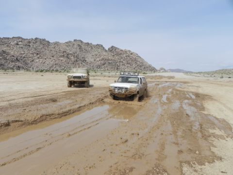 1978 Ford Bronco - Two off-road vehicles navigate a challenging muddy track under a clear sky.