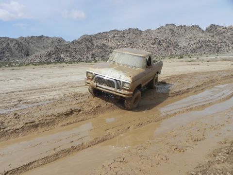 1978 Ford Bronco - A tan Ford F-Series truck navigates a challenging muddy terrain under a bright sky with rocky hills in the background.