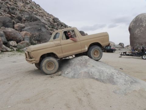 1978 Ford Bronco - A tan 1978 Ford Bronco navigates rocky terrain off-road. Other vehicles and a trailer are visible in the background.