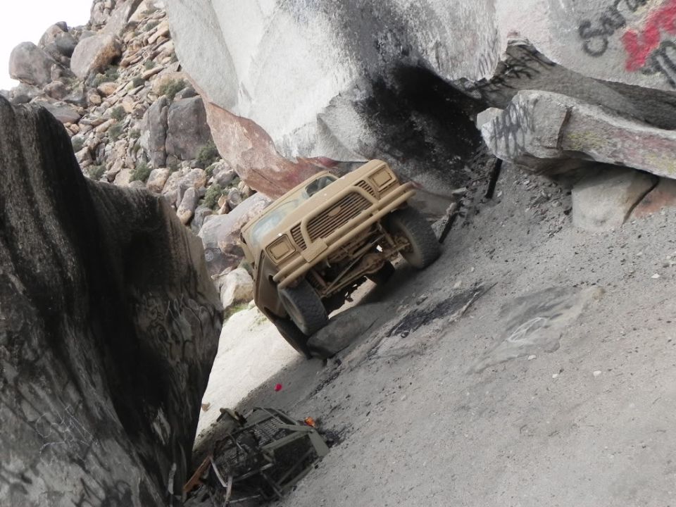 Tan off-road vehicle climbing a steep, rocky incline next to large boulders. Another vehicle is visible below