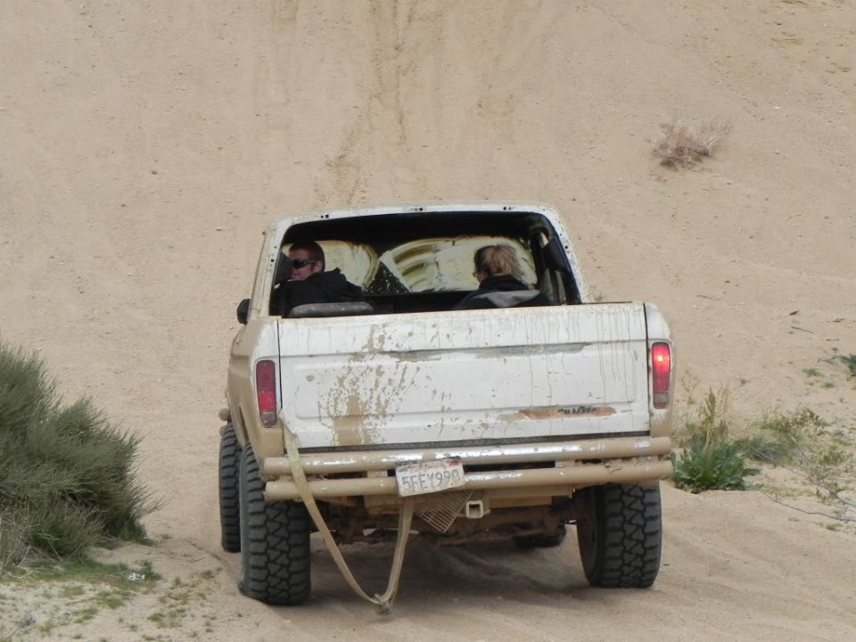 Rear view of a white 1978 Ford F-150 pickup truck driving on a dirt road, two people inside