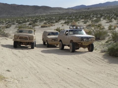 1978 Ford Bronco - Three off-road vehicles traverse a dusty desert trail under a clear sky.