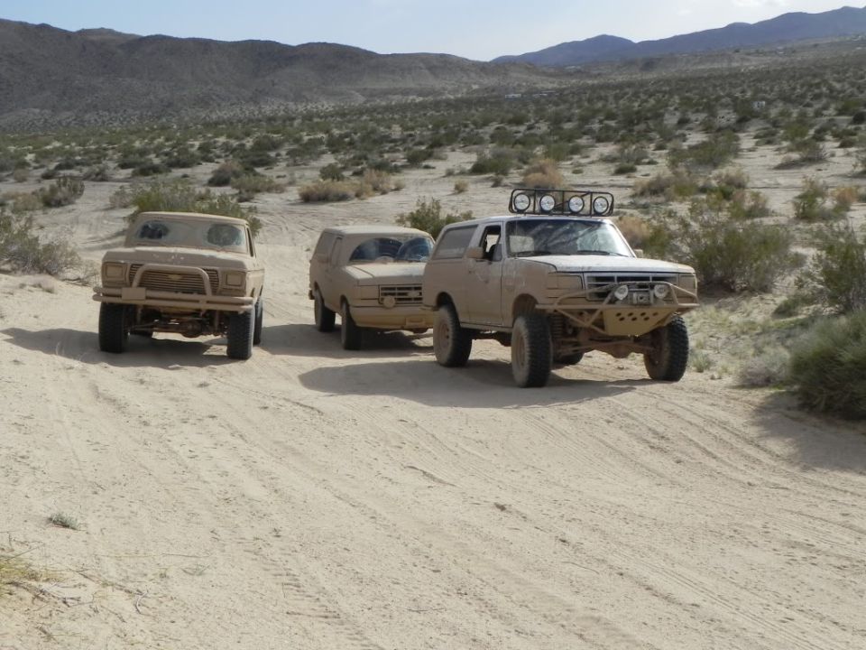 Three tan off-road vehicles on a dirt road, one with a light bar on top