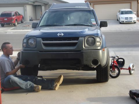 2004 Nissan Xterra - A man works on the front bumper of a blue 2004 Nissan Xterra, with a red tricycle parked beside it.