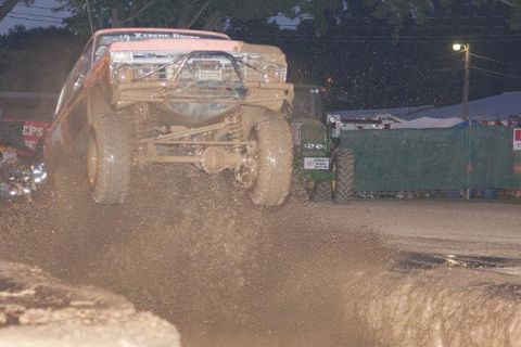 1978 Ford Bronco - A modified truck jumps through a mud pit, spraying dirt and water, with a tractor visible behind a green barrier.