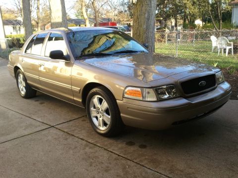 2007 Ford Crown Victoria - A tan 2007 Ford Crown Victoria is parked in front of a residential yard with trees and chairs visible.
