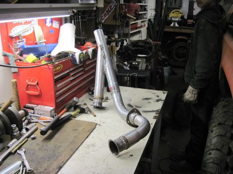 1978 Ford Bronco - A custom exhaust pipe is laid out on a workbench in a garage setting, surrounded by tools and equipment.