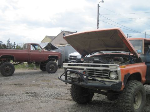 1978 Ford Bronco - Two lifted pickup trucks are parked outdoors, one with its hood open, revealing the engine.