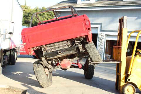 1978 Ford Bronco - A red pickup truck is precariously balanced on forklift forks, with another truck visible in the background.