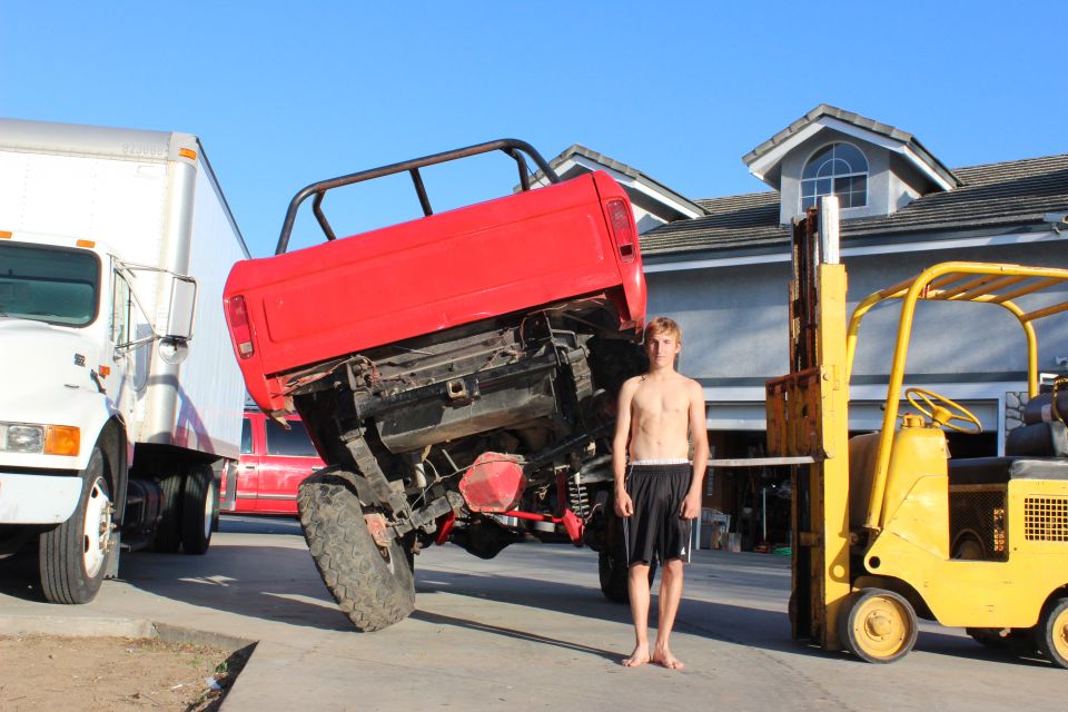 Red truck bed tilted up, person standing beside it. White truck and yellow forklift visible