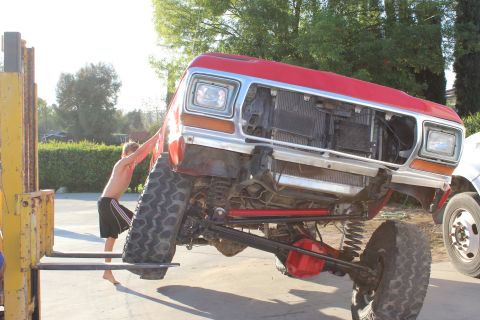 1978 Ford Bronco - A red pickup truck is being lifted by a forklift, with a person assisting. Another vehicle is visible in the background.