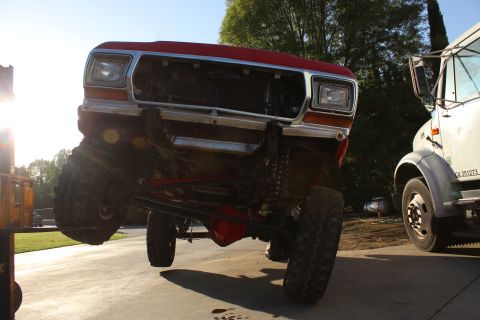 1978 Ford Bronco - A red Ford Bronco is lifted by a forklift, showing its front end and suspension modifications, with a white truck nearby.