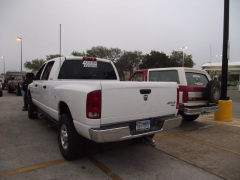 1989 Ford Bronco - A red and white SUV is parked behind a white pickup truck, showing its rear profile and spare tire.