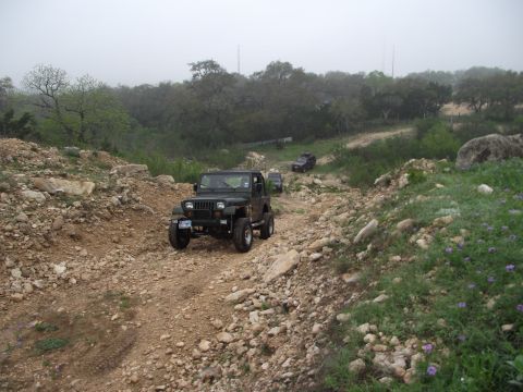 1989 Ford Bronco - Several Jeeps navigate a , dusty trail surrounded by trees and rocks under a hazy sky.