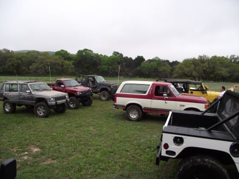 1989 Ford Bronco - A collection of modified SUVs and trucks are gathered outdoors on a cloudy day.