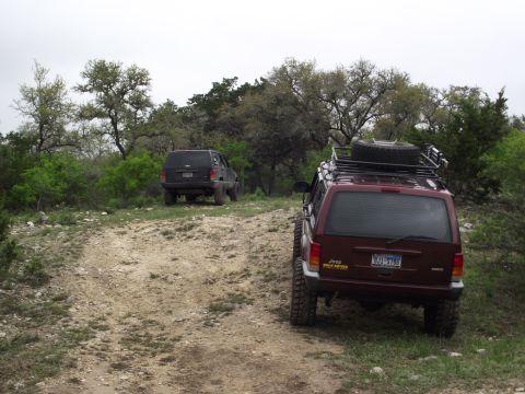 1989 Ford Bronco - Two Jeep Cherokee XJs navigate a dirt path through a wooded, hilly area.