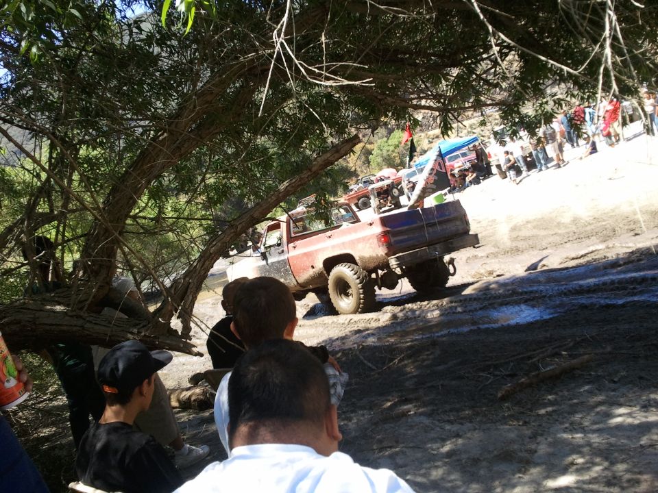 Red and blue pickup truck with large tires on a muddy trail, rear view