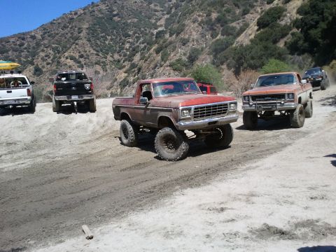 1978 Ford Bronco - A red lifted pickup truck navigates a dirt road alongside other off-road vehicles.
