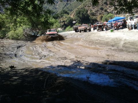 1978 Ford Bronco - Multiple off-road vehicles navigate a muddy terrain with trees and hills in the background.
