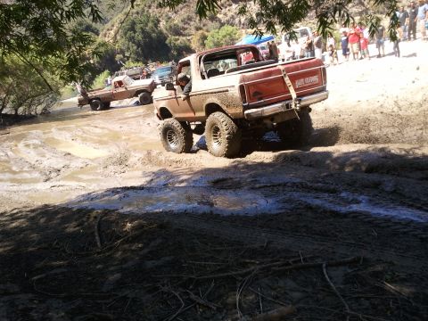 1978 Ford Bronco - Several off-road vehicles navigate a muddy terrain with spectators watching from a distance.