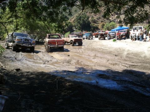 1978 Ford Bronco - Several trucks and SUVs navigate a muddy off-road course under trees and hills.