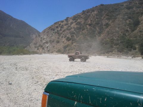 1978 Ford Bronco - Off-road trucks navigate a dusty dry riverbed with steep, vegetated hillsides under a clear blue sky.