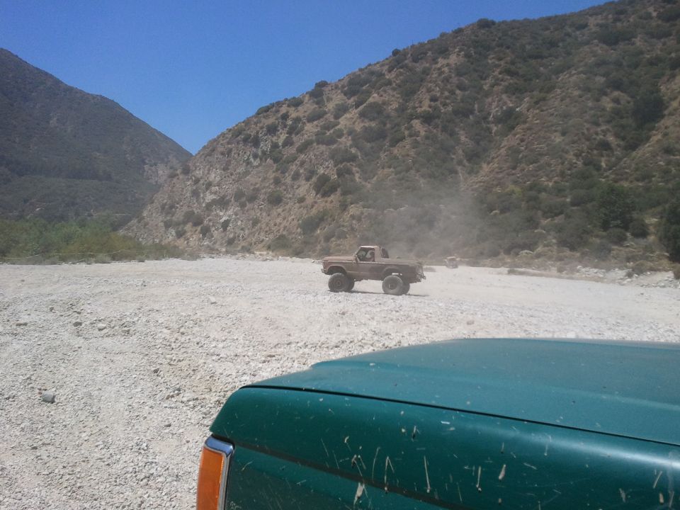 Two trucks driving off-road in a dry riverbed with mountains in the background. One truck is in the foreground, partially visible